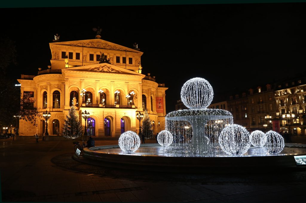 Gebäudefront der Alten Oper in Frankfurt bei Nacht mit einem Brunnen geschmückt von Kugeln aus Lichterketten im Vordergrund.