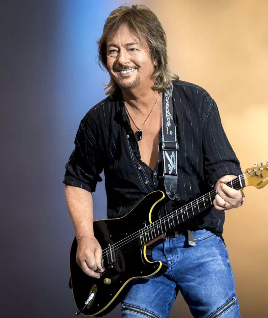 Chris Norman steht mit einer Gitarre und lächelt nach vorne.