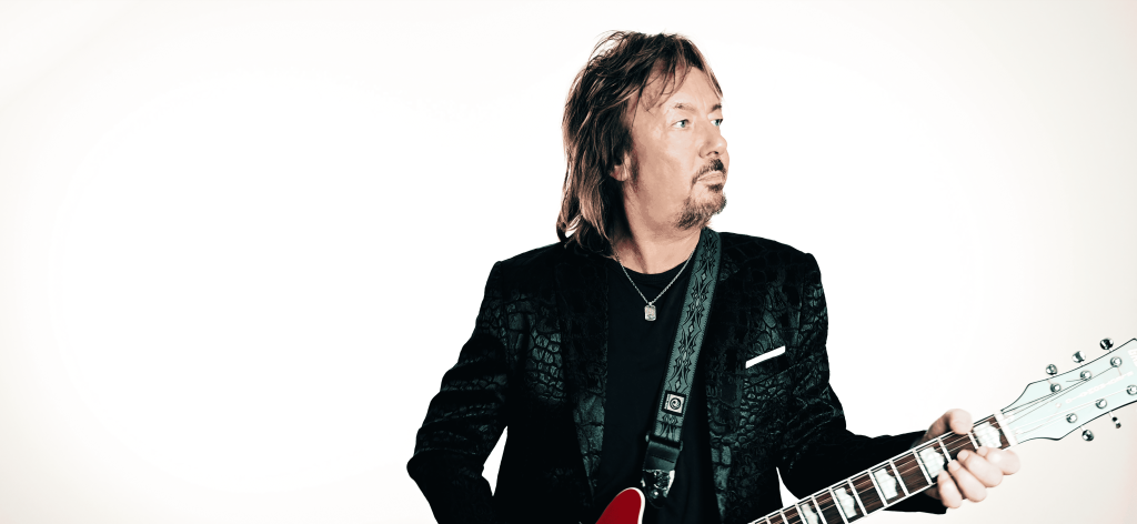 Chris Norman steht mit einer Gitarre vor einem weißen Hintergrund.